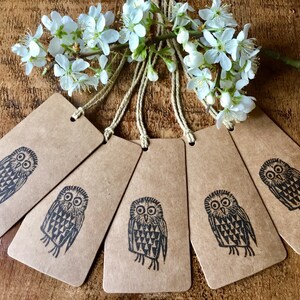 May include: Six brown paper tags with a black owl illustration and string. The tags are arranged in a fan shape, with white flowers and a wooden background.