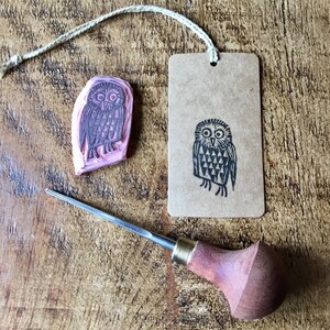 May include: A linocut owl stamp with a matching print on a tag, and a wood carving tool. The stamp is pink with a dark grey owl design. The tag is brown with a black owl print. The wood carving tool has a wooden handle and a metal blade.