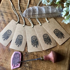 May include: Several brown paper gift tags, each featuring a black owl print, are displayed with twine. A carved owl stamp and a wood carving tool are also visible. A large feather and white flowers are in the background.