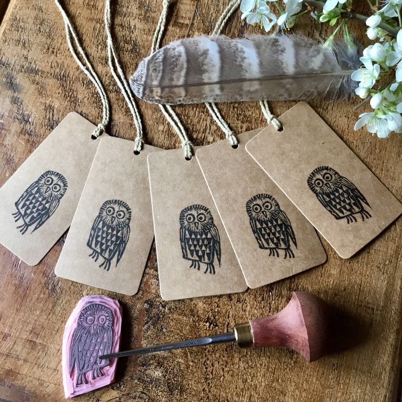 May include: Several brown paper gift tags, each featuring a black owl print, are displayed with twine. A carved owl stamp and a wood carving tool are also visible. A large feather and white flowers are in the background.