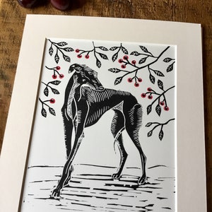 May include: Black and white linocut print of a greyhound dog beneath cherry tree branches with red cherries. The print is matted in a cream-colored mat. The title "Beneath the Cherry Tree" is printed at the bottom.