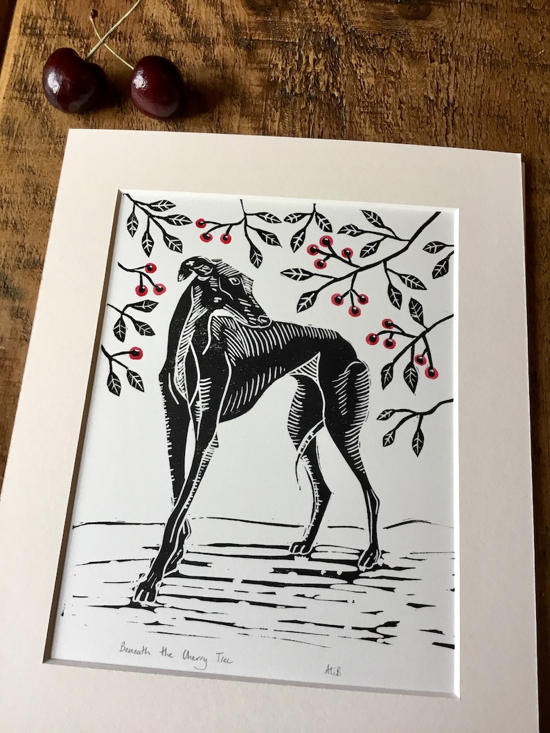 May include: Black and white linocut print of a greyhound dog beneath cherry tree branches with red cherries. The print is matted in a cream-colored mat. The title "Beneath the Cherry Tree" is printed at the bottom.