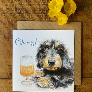 May include: A greeting card featuring a black and tan dog with a glass of beer. The card says "Cheers!" in blue lettering.