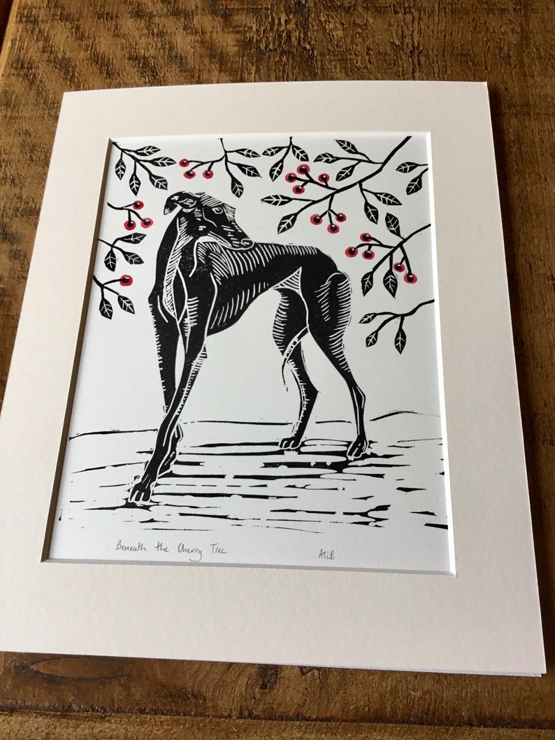 May include: Black and white linocut print of a greyhound dog beneath a cherry tree. The dog is the focal point, with detailed linework. Red cherries and green leaves are scattered throughout the branches. The print is matted in a cream-colored mat.