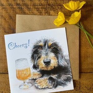 May include: A greeting card with a black and brown dog with a glass of beer. The card says "Cheers!" in blue text.