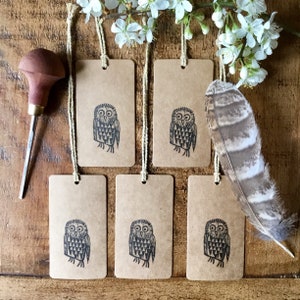 May include: Rectangular kraft paper gift tags featuring a black owl print, strung with twine. A wooden carving tool and a feather are also visible. White flowers are in the background. The tags have a rustic appearance.