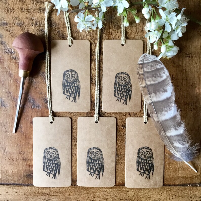 May include: Rectangular kraft paper gift tags featuring a black owl print, strung with twine. A wooden carving tool and a feather are also visible. White flowers are in the background. The tags have a rustic appearance.