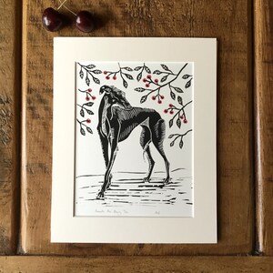 May include: Black and white linocut print of a greyhound dog standing beneath a cherry tree with red cherries. The print is matted in a cream-colored mat and framed. Two cherries rest on the wooden surface.