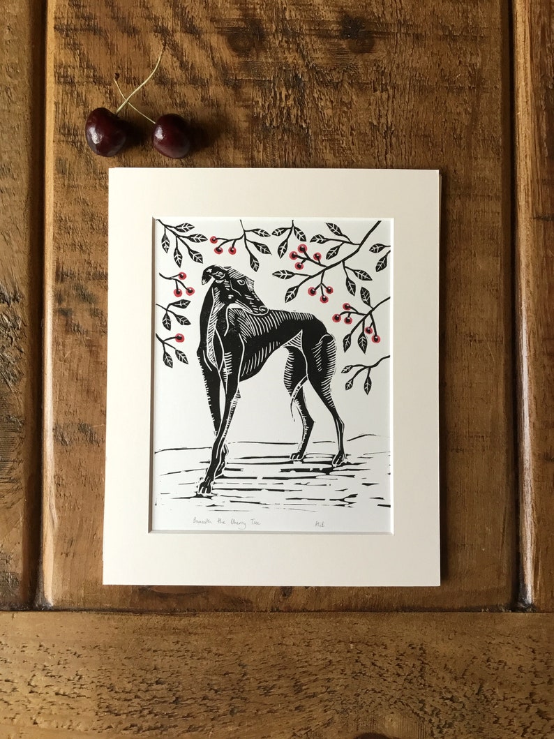 May include: Black and white linocut print of a greyhound dog standing beneath a cherry tree with red cherries. The print is matted in a cream-colored mat and framed. Two cherries rest on the wooden surface.
