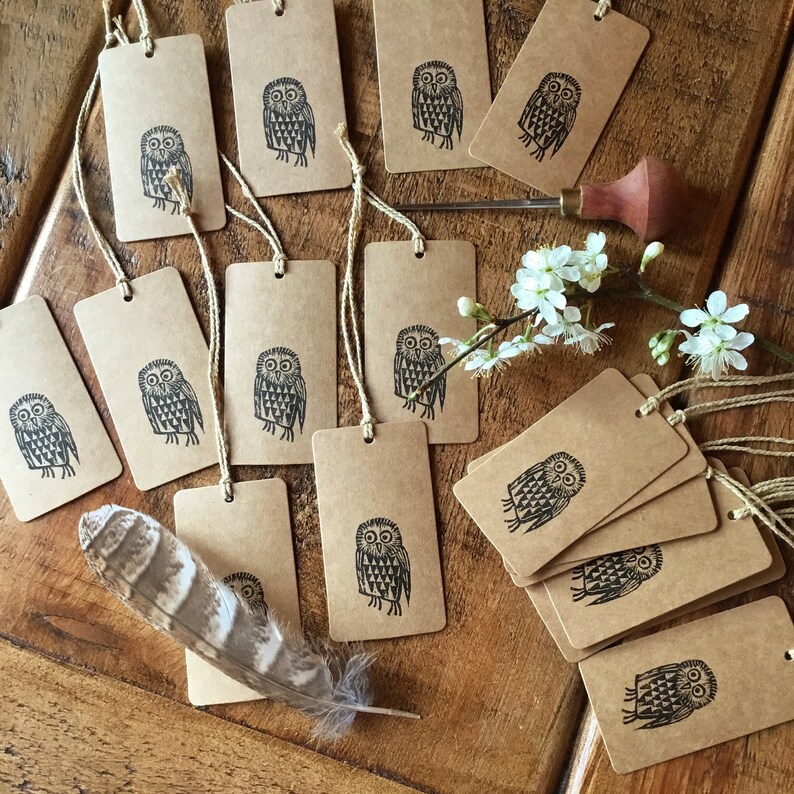 May include: Kraft paper gift tags with a black owl design and twine. The tags are rectangular with rounded corners. A feather and a carving tool are also visible.
