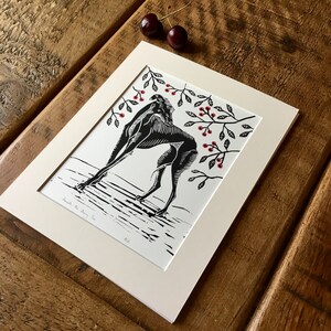 May include: Black and white linocut print titled "Beneath the Cherry Tree" featuring a stylized figure with cherry branch accents. The print is matted in a cream-colored mat and framed. Two cherries rest on the wooden surface.