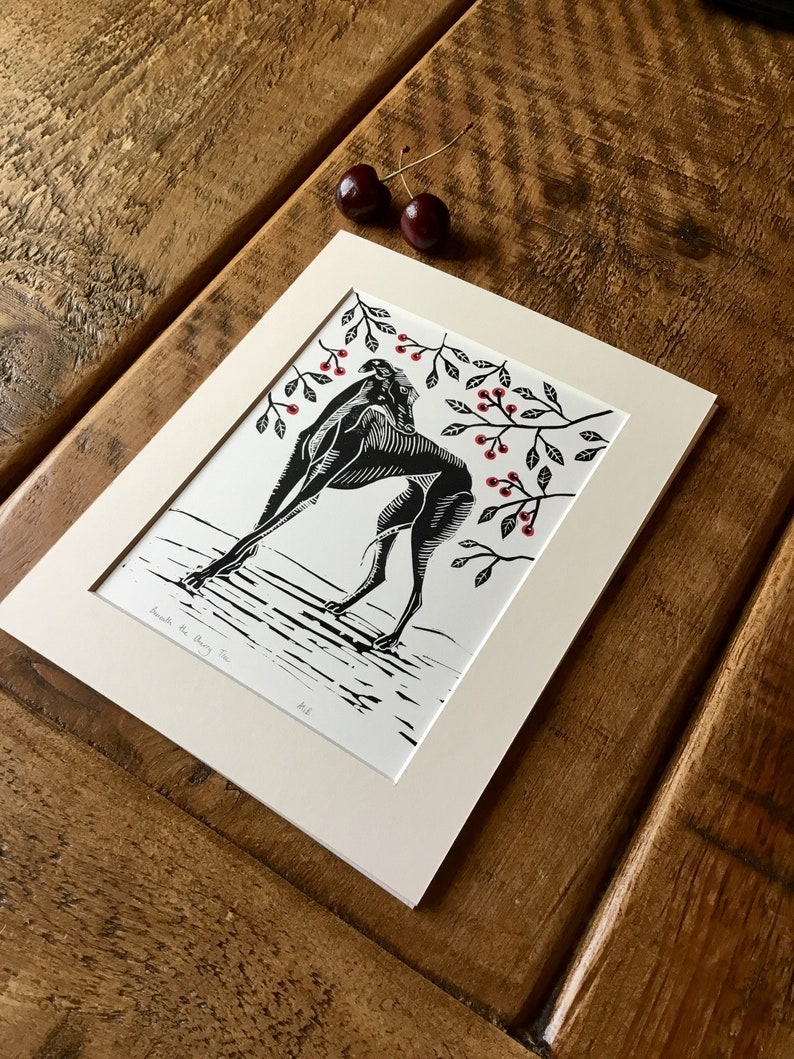 May include: Black and white linocut print titled "Beneath the Cherry Tree" featuring a stylized figure with cherry branch accents. The print is matted in a cream-colored mat and framed. Two cherries rest on the wooden surface.