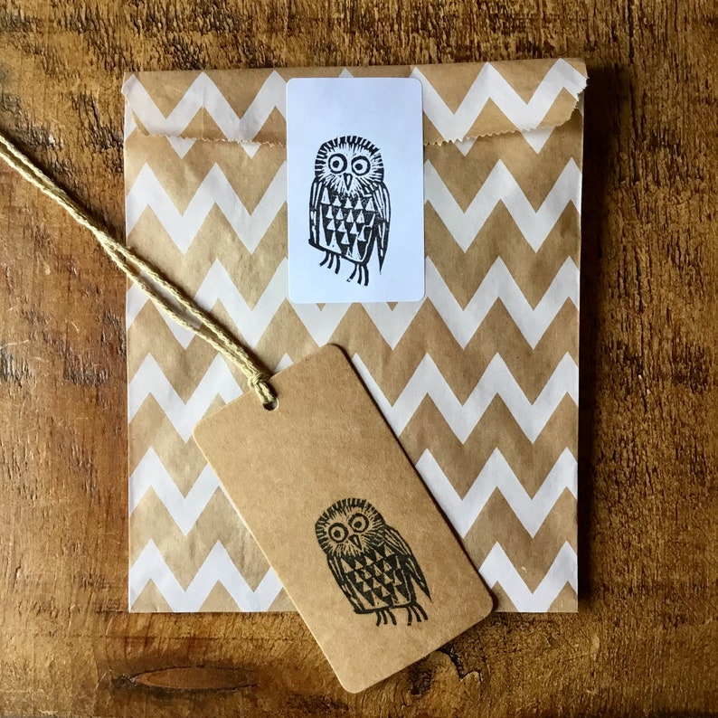 May include: A brown paper bag with a white zig-zag pattern, a white rectangular sticker, and a brown tag. Each features a black and white owl illustration. A natural twine string is attached to the tag.