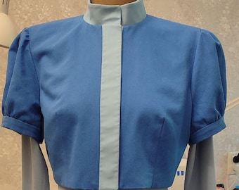 May include: A two-piece dress on a mannequin. The top is a light blue, short-sleeved bolero with a high collar and a light gray center panel. The bottom is a light gray, long-sleeved sheath dress.