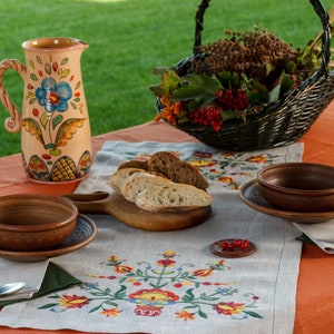 May include: An outdoor dining scene with a ceramic jug decorated with flowers, bread slices on a wooden board, and two bowls. A woven basket contains berries and foliage. A linen tablecloth with embroidered flowers is also present.
