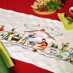 May include: White embroidered table runner with a nativity scene design. The scene includes a manger, baby Jesus, Mary, Joseph, angels, sheep, and a star. The runner is perfect for a Christmas table setting.