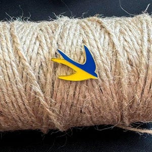 May include: A gold-colored pin with a blue and yellow stylized bird design. The pin is on a brown rope background.