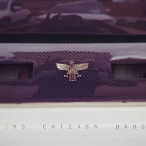 Puede incluir: Primer plano de un emblema de águila plateado en un coche negro y blanco. El emblema es un águila estilizada con las alas extendidas y una corona en la cabeza. El coche tiene un acabado de fibra de carbono y el texto "Screaming Chicken Badge" está escrito debajo del emblema.