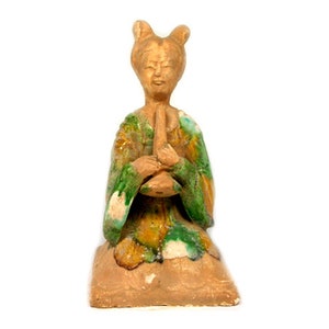 May include: A ceramic figurine of a woman playing a flute, wearing a green and yellow robe. The figurine is sitting on a brown base.
