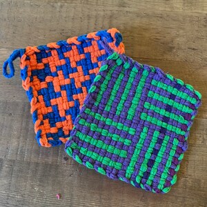 Może przedstawiać: Dwa tkane uchwyty do garnków, jeden w kratkę niebiesko-pomarańczową, a drugi w kratkę zielono-fioletową.