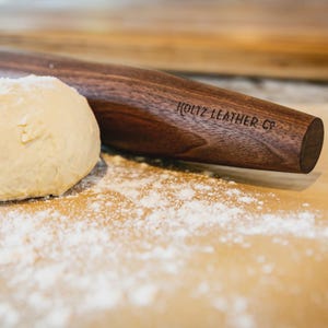 May include: A dark brown wooden rolling pin with the text KOLTZ LEATHER CO engraved on the side, positioned next to a round ball of beige dough on a surface dusted with white flour. The rolling pin has a smooth, cylindrical wooden finish.