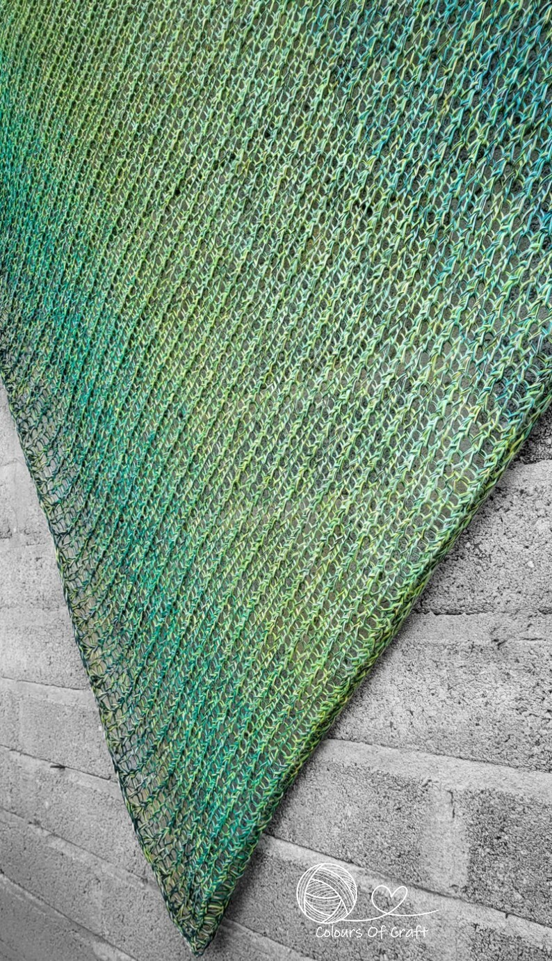 Op de afbeelding: Een groene en blauwe gebreide sjaal met een verloopmotief. De sjaal is over een grijze bakstenen muur gelegd.