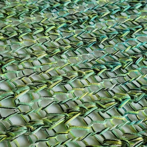 Op de afbeelding: Een close-up van een groene, blauwe en gele gehaakte stof met een zwarte draad die er doorheen geweven is. De stof is gemaakt met een herhalend patroon van knopen en lussen.