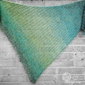 Op de afbeelding: Een groene en blauwe ombre gebreide sjaal met een diagonaal patroon. De sjaal hangt aan een bakstenen muur.
