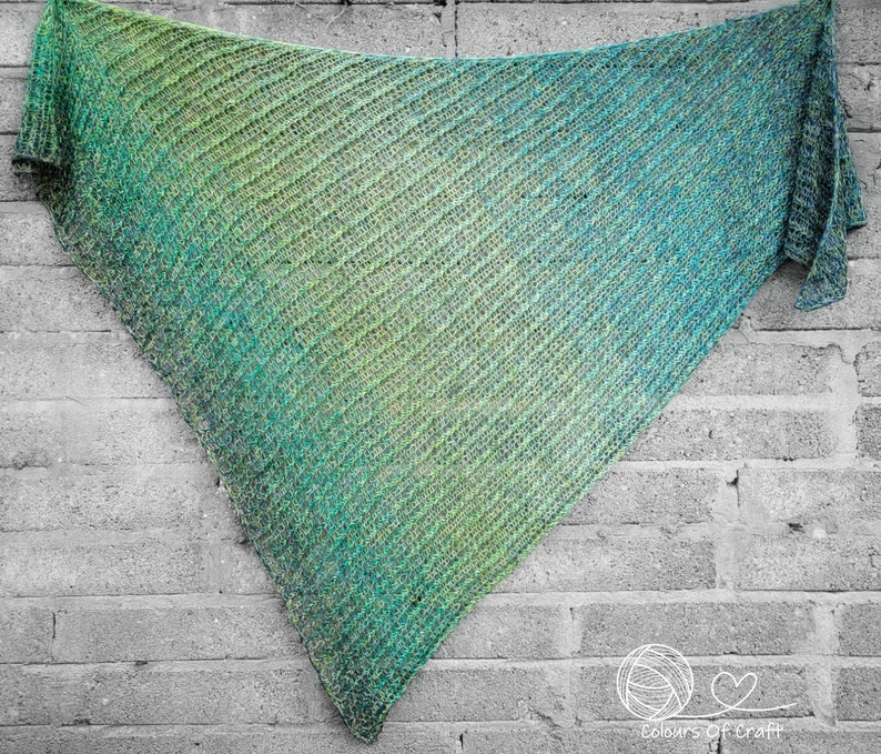 Op de afbeelding: Een groene en blauwe ombre gebreide sjaal met een diagonaal patroon. De sjaal hangt aan een bakstenen muur.