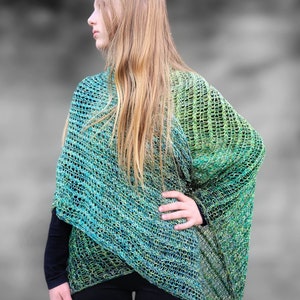 Op de afbeelding: Een groene en blauwe ombre gebreide sjaal met een losse, open structuur. De sjaal is over de schouders van een persoon gedrapeerd en wordt gedragen met een zwarte broek.