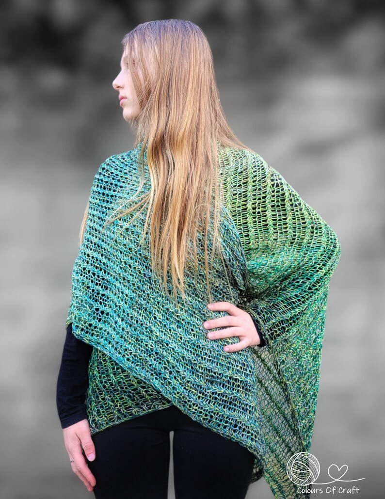Op de afbeelding: Een groene en blauwe ombre gebreide sjaal met een losse, open structuur. De sjaal is over de schouders van een persoon gedrapeerd en wordt gedragen met een zwarte broek.
