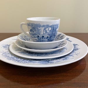 May include: A white ceramic teacup and a stack of three plates of varying sizes, all featuring a blue transferware pattern depicting a landscape with a stone bridge, trees, and buildings. The pieces are arranged in a vertical stack.