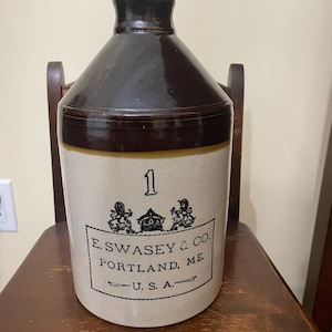 May include: A stoneware jug with a dark brown glazed neck and a cream-coloured body. The front features the number 1, a crown emblem, and the text E. SWASEY & CO. PORTLAND, ME. U. S. A. printed in black ink within a rectangular border.