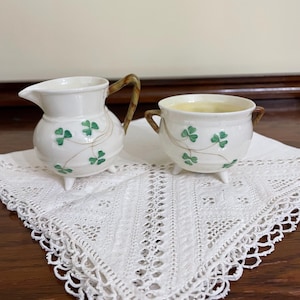 May include: A cream-coloured ceramic creamer and sugar bowl set featuring a hand-painted green shamrock pattern with thin brown stems. Both pieces have three small feet, and the creamer includes a curved brown handle, while the sugar bowl has two small brown side handles.