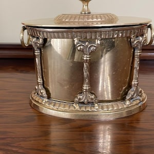 Peut inclure: Un récipient cylindrique en laiton avec un couvercle amovible doté d'un fleuron central. Le corps est orné de colonnes décoratives verticales et de deux poignées latérales en anneau. La base et les bords du couvercle présentent une bordure festonnée.