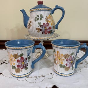 May include: A ceramic teapot and two matching mugs featuring a cream-coloured base with vertical ridges, blue rims, and blue handles. The pieces are decorated with hand-painted floral motifs in shades of red, yellow, and green, with small brown speckles.
