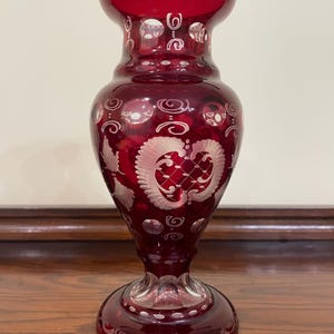 May include: A deep red glass vase featuring intricate white etched patterns, including floral motifs, lattice designs, and circular accents. The vase has a flared rim, a tapered neck, a rounded body, and a circular base with clear glass detailing.