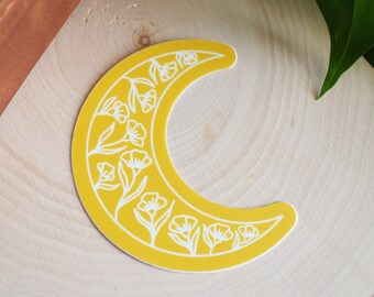 May include: A yellow crescent moon sticker with white floral designs. The sticker is on a light wood surface.
