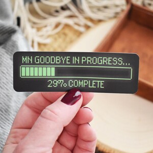 May include: A black and green sticker with the text "MN GOODBYE IN PROGRESS..." and a progress bar that is 29% complete. The sticker is being held by a hand.