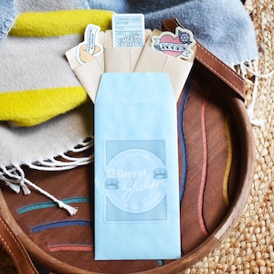 May include: A light blue envelope with the text "12 Days of Stickers" sits in a wooden tray with a leather handle. Several small envelopes with stickers are visible, along with a folded blanket.
