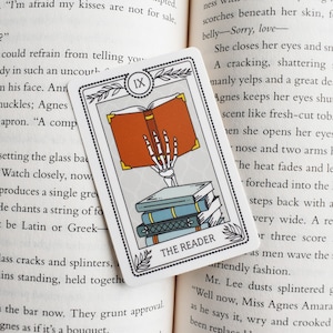 May include: A tarot card with a skeletal hand holding a red book. The card is titled "The Reader" and has the Roman numeral IX in the top right corner. The card is on a book with text about a woman named Agnes.