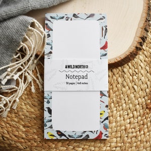Puede incluir: Un bloc de notas con fondo blanco y un borde azul con un patr&oacute;n de p&aacute;jaros coloridos. El bloc de notas tiene 50 p&aacute;ginas y mide 10,2 x 20,3 cm. El texto "WILD NORTH CO" y "Notepad" est&aacute; impreso en la parte delantera.