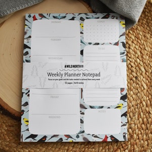 Puede incluir: Un bloc de notas azul con un patr&oacute;n de p&aacute;jaros y el texto "#WILD NORTH CO Weekly Planner Notepad Focus on your goals and the tasks needed to achieve them every week 52 pages | 8x11 inches"