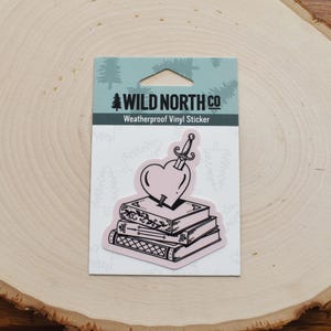 May include: A black and white sticker featuring a heart pierced by a dagger, sitting on top of a stack of books. The sticker is labeled "Wild North Co. Weatherproof Vinyl Sticker".