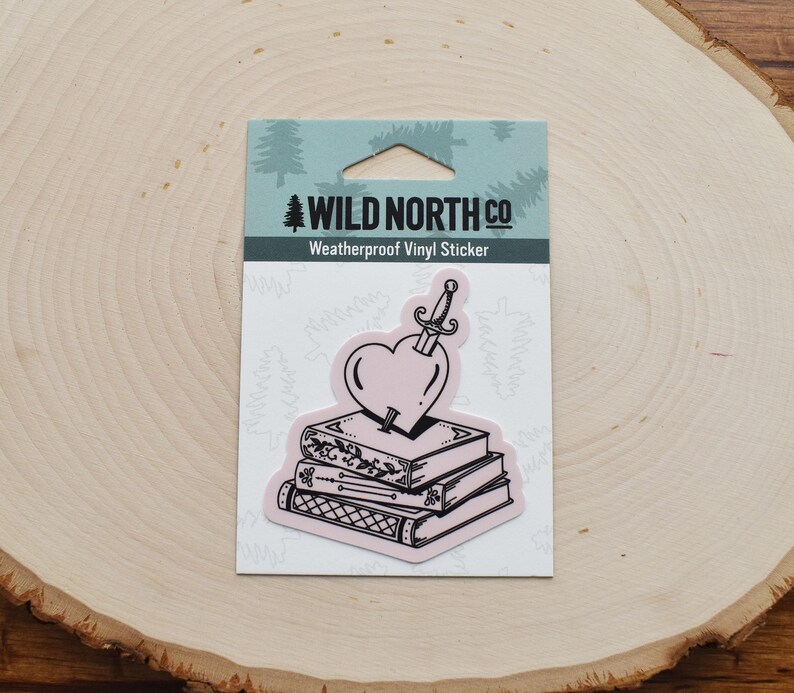 May include: A black and white sticker featuring a heart pierced by a dagger, sitting on top of a stack of books. The sticker is labeled "Wild North Co. Weatherproof Vinyl Sticker".