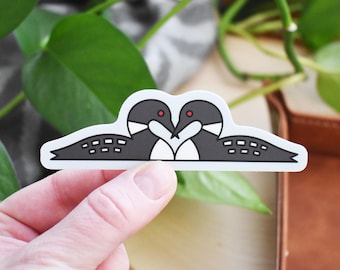 Può includere: Un adesivo raffigurante due strolaghe stilizzate, con i becchi che si toccano a formare un cuore. Le strolaghe sono nere e bianche con occhi rossi, su sfondo bianco. L'adesivo è tenuto in mano.