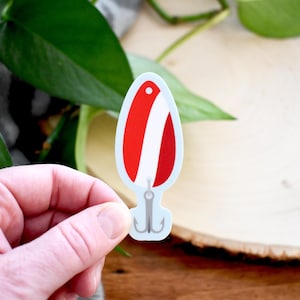 Puede incluir: Una pegatina de señuelo de pesca con un diseño rojo y blanco. La pegatina tiene un borde azul claro y un anzuelo plateado en la parte inferior. El señuelo se sostiene en la mano de una persona.