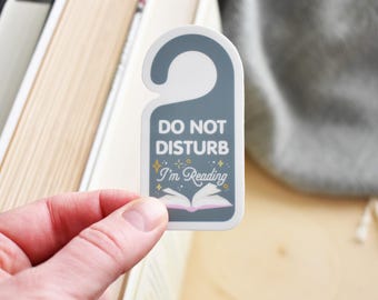 May include: A gray door hanger with the text "Do Not Disturb I'm Reading" and a pink and white open book illustration. The hanger has a white hook at the top.