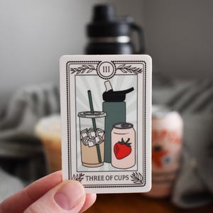 May include: A rectangular sticker featuring a tarot card design with the text "THREE OF CUPS." The card depicts a clear iced coffee, a water bottle, and a can with a strawberry. The card has a white border and a light green background.