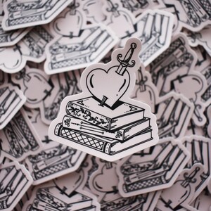 May include: A black and white illustration of a stack of books with a heart pierced by a dagger on top. The heart is surrounded by a white background.
